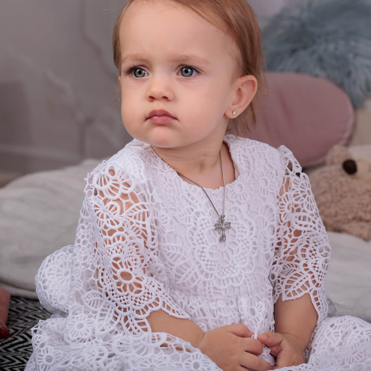 Child wearing a Christening Gown for Girls with a cross necklace, sitting on a soft surface.