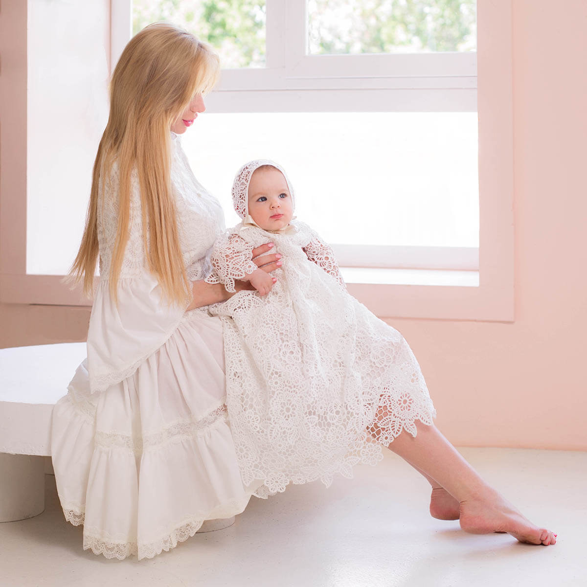 Woman in a Christening Gown for Girls holding a baby in a similar outfit by a window.