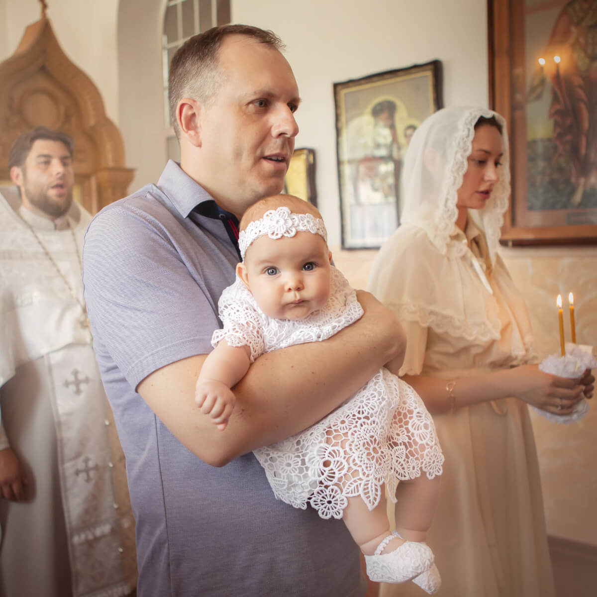 Man holding a baby in a Lace Baptism Dress with a woman in a headscarf and candle in a church setting.