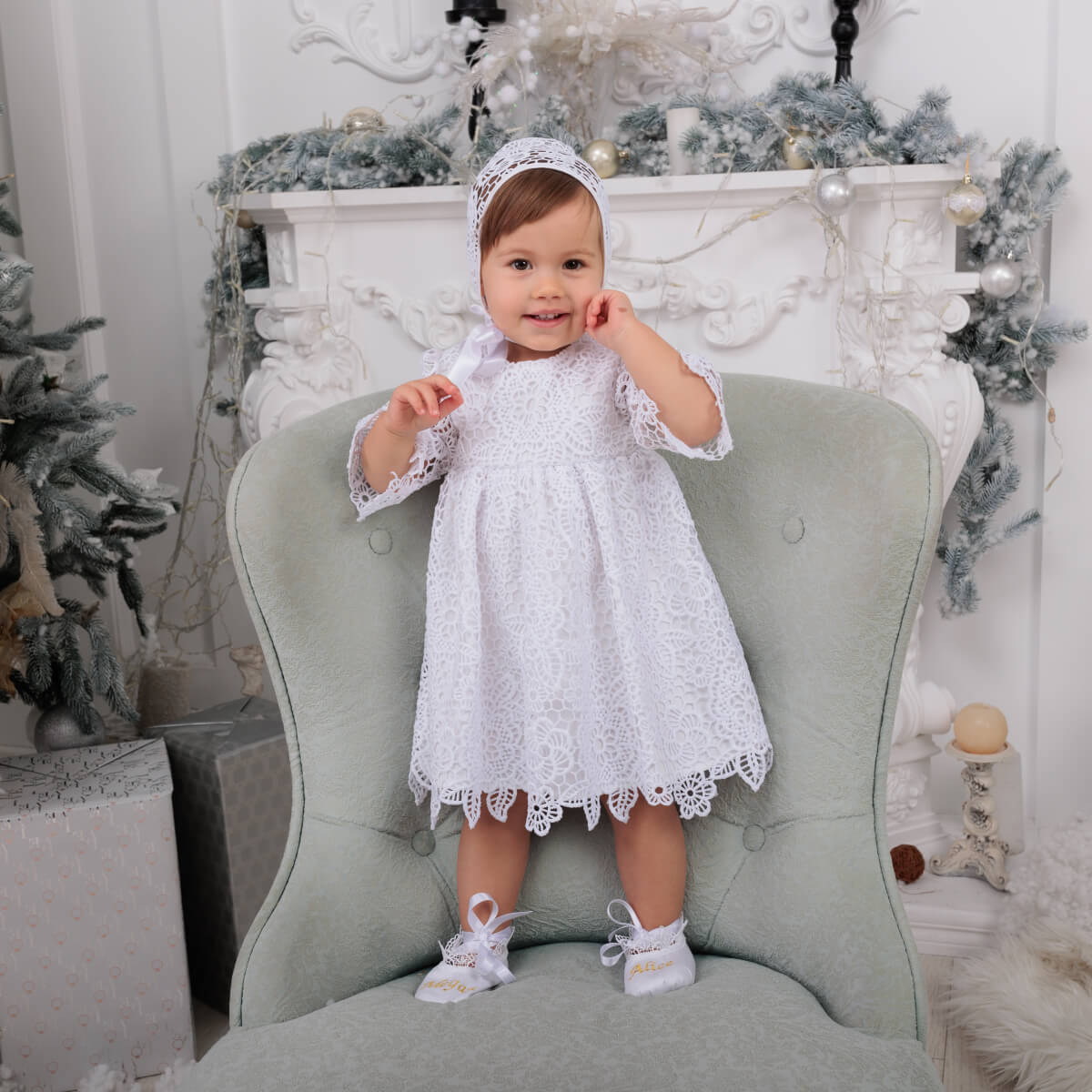 Baby in a Lace Baptism Dress sitting on a green chair with Christmas decorations in the background