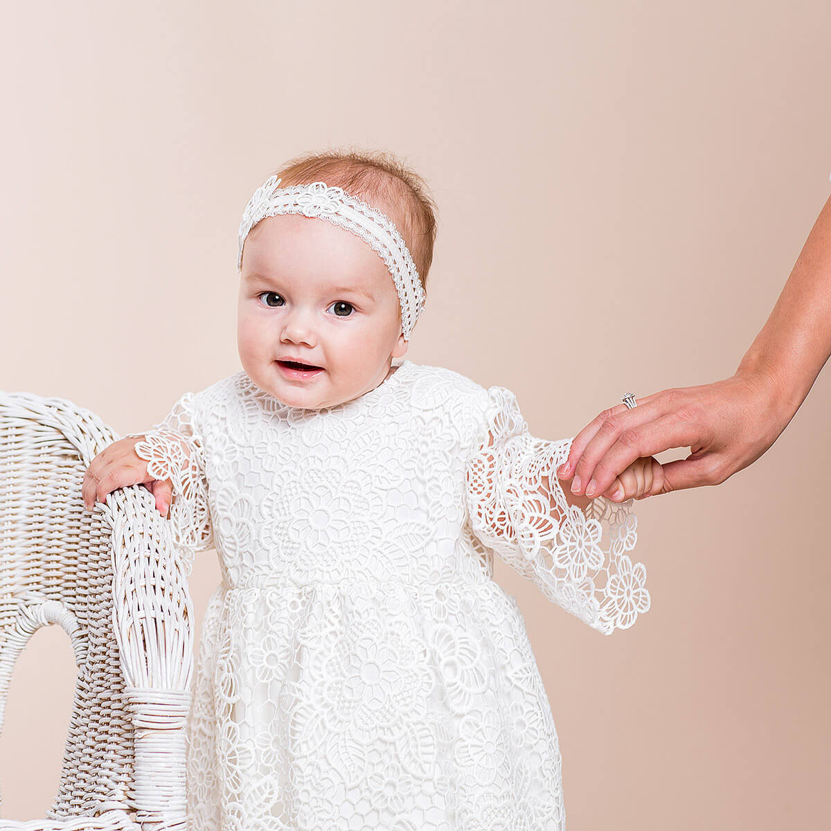 Baby in a Baby Baptism Dress with a headband, standing next to a wicker chair on a beige background.