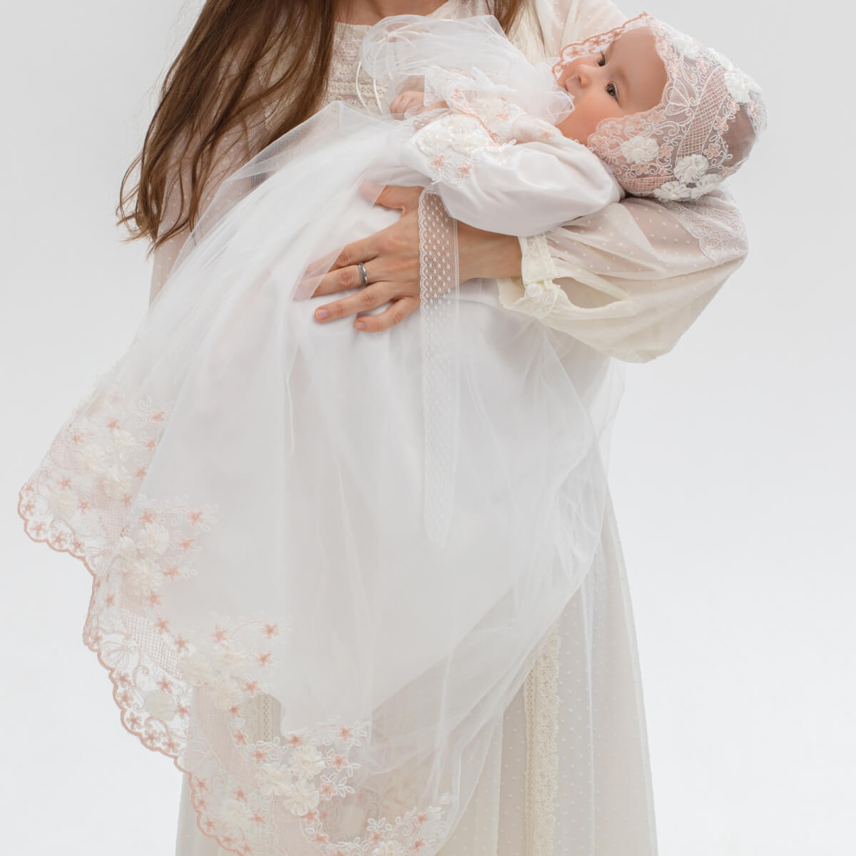 Woman holding a baby wrapped in a flower lace baptism bonnet against a white background