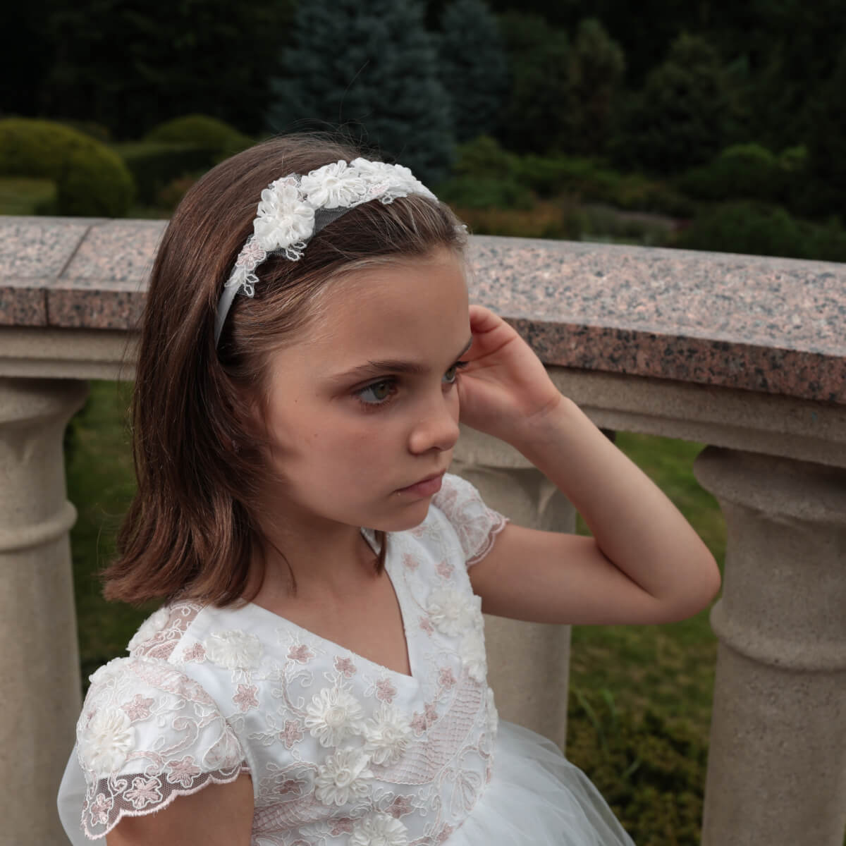 Young girl in a white lace dress with a headband, standing outdoors.
