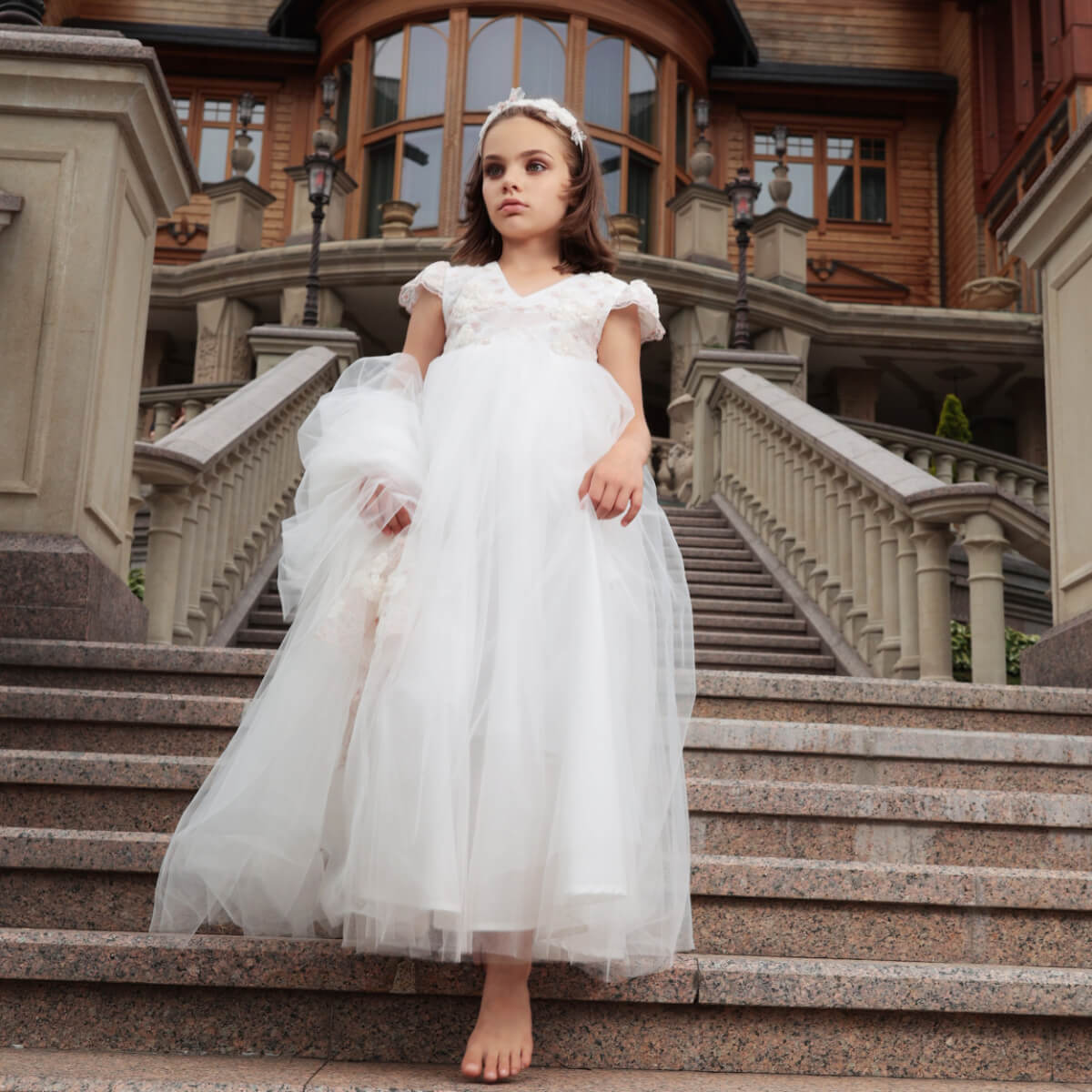 Young girl in lace flower girl dress with train standing on stone steps in front of an elegant building.