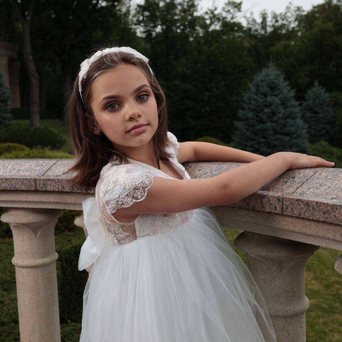 Young girl in flower girl dress with train standing outdoors on a stone bench.