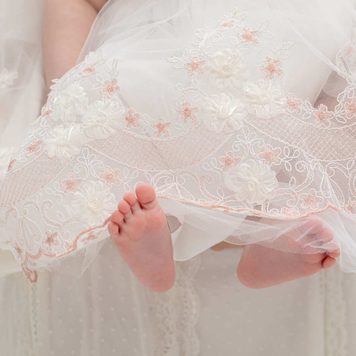 Close-up of a baby's feet peeking out from under a Baptism Dress with Floral Lace with floral details.
