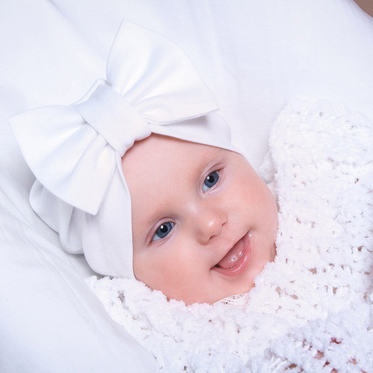 Baby wearing a white baby turban with a large bow, surrounded by soft white fabric.