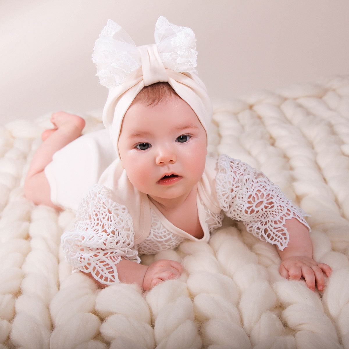 Baby in a white lace outfit and headband sitting on a white textured surface