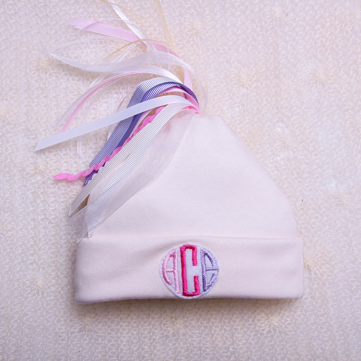 White baby hat with pink and purple monogram on a textured white background
