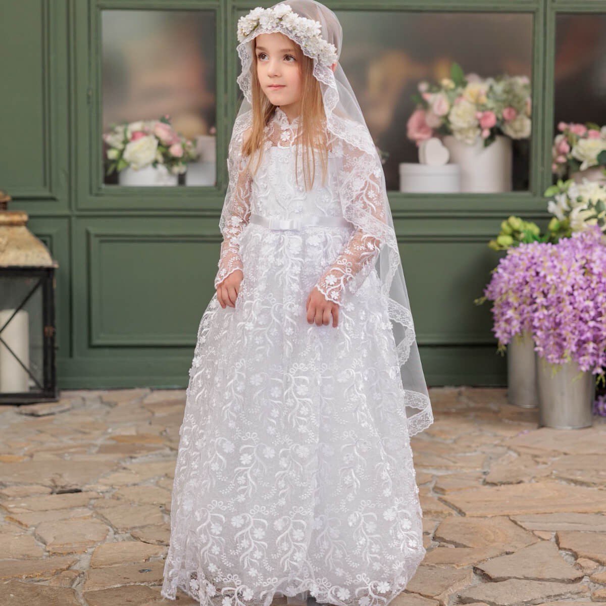 Young girl in a white lace dress standing outdoors with flowers and a green wall in the background.