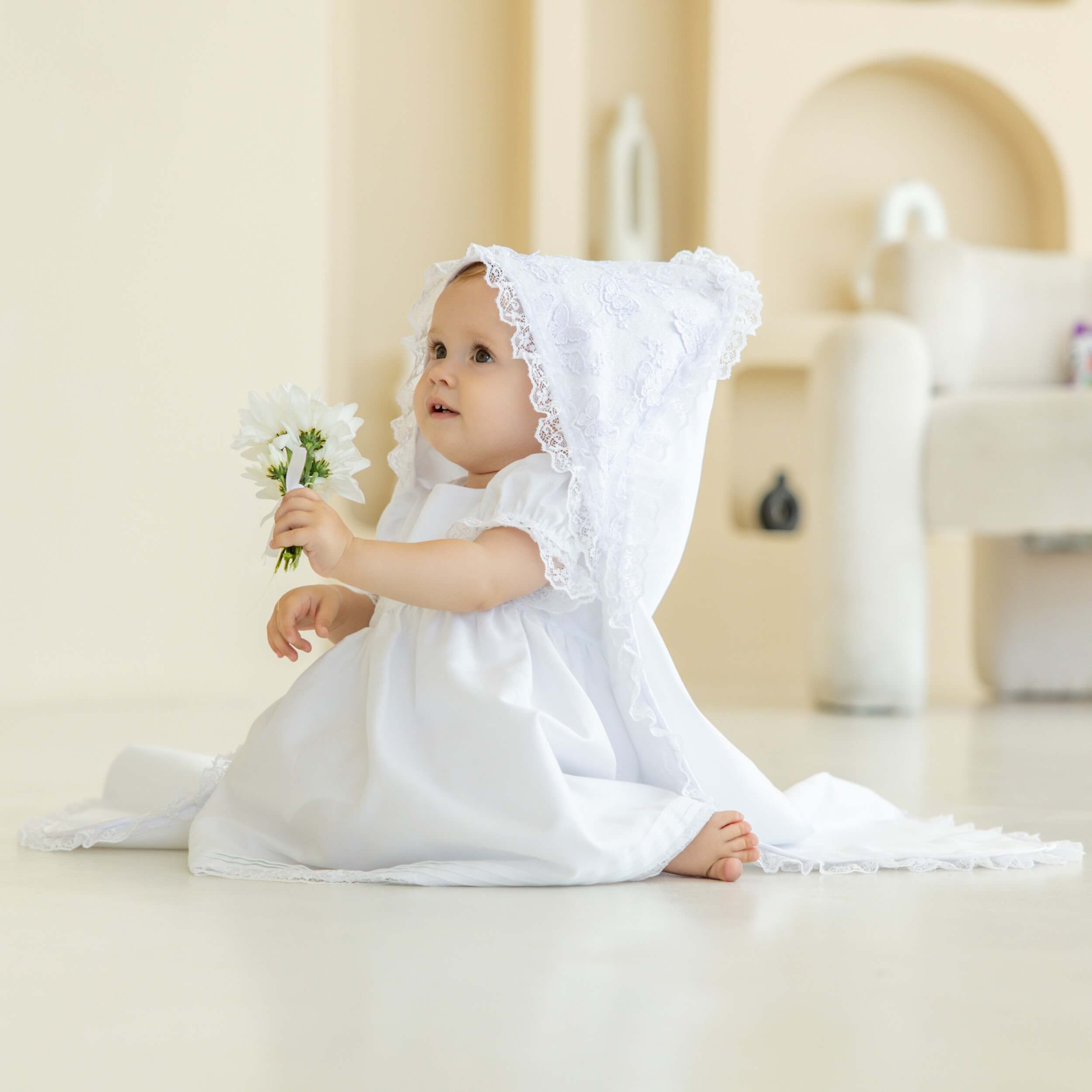 Baby in a white dress with a hooded christening blanket sitting on a light-colored floor.