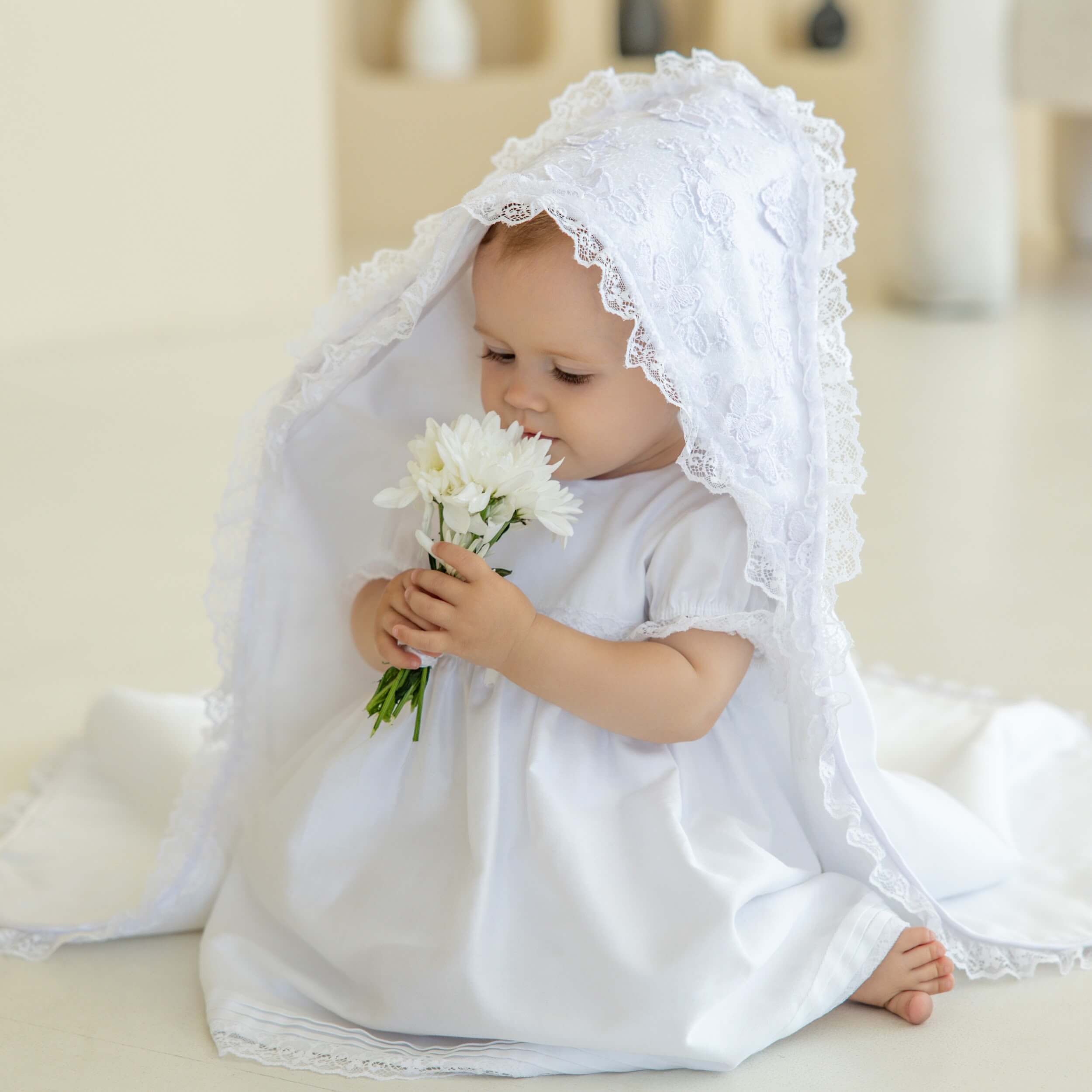 Baby in a white dress with a christening blanket holding flowers indoors
