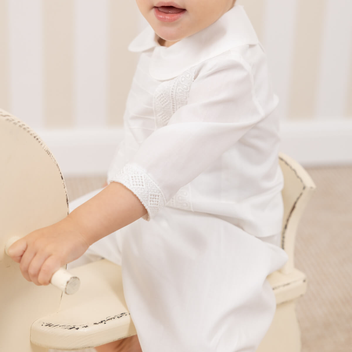 Child wearing a Baptism Gown for Boy sitting on a beige chair.