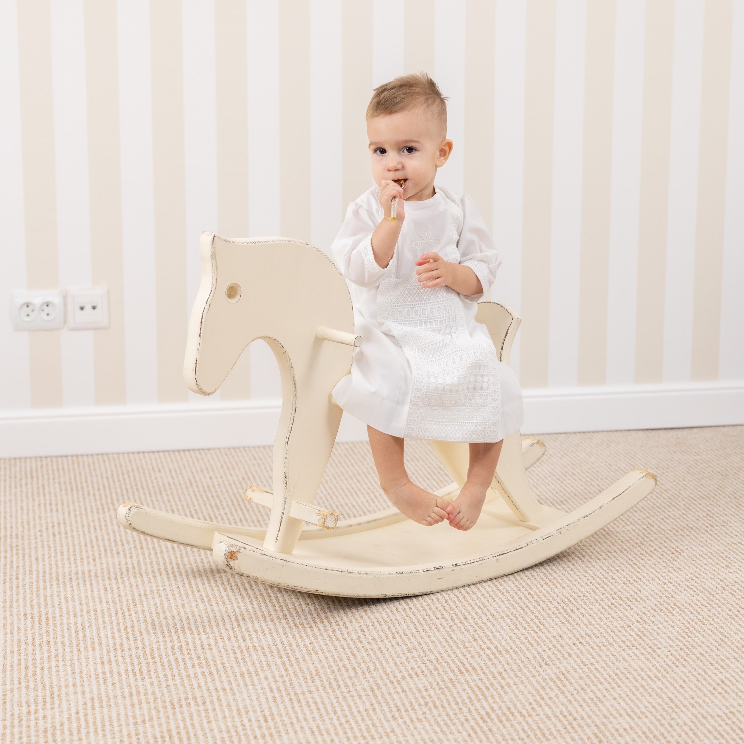 Baby sitting on a white rocking horse in Baptism Gown for Boy a room with light-colored walls and carpet.