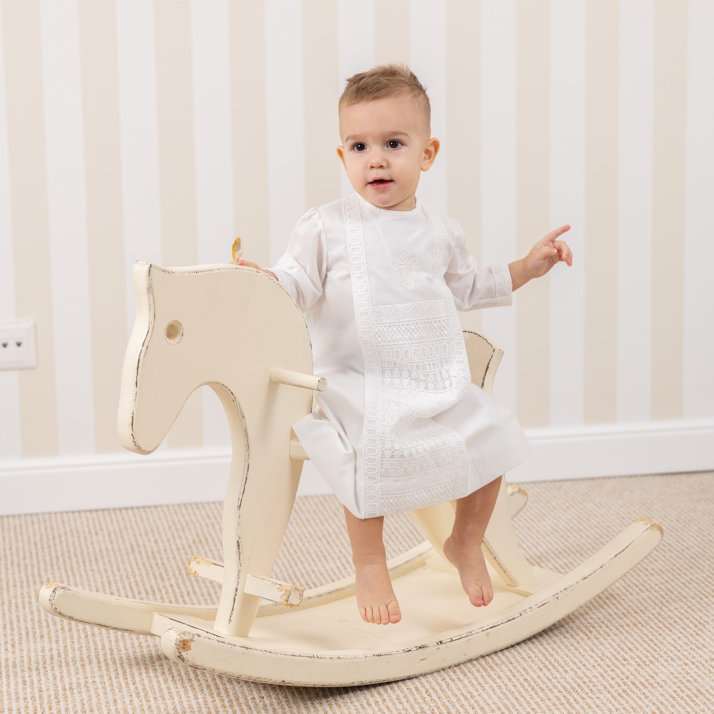 Child in a  Baptism Gown for Boy sitting on a wooden rocking horse in a room with striped wallpaper.