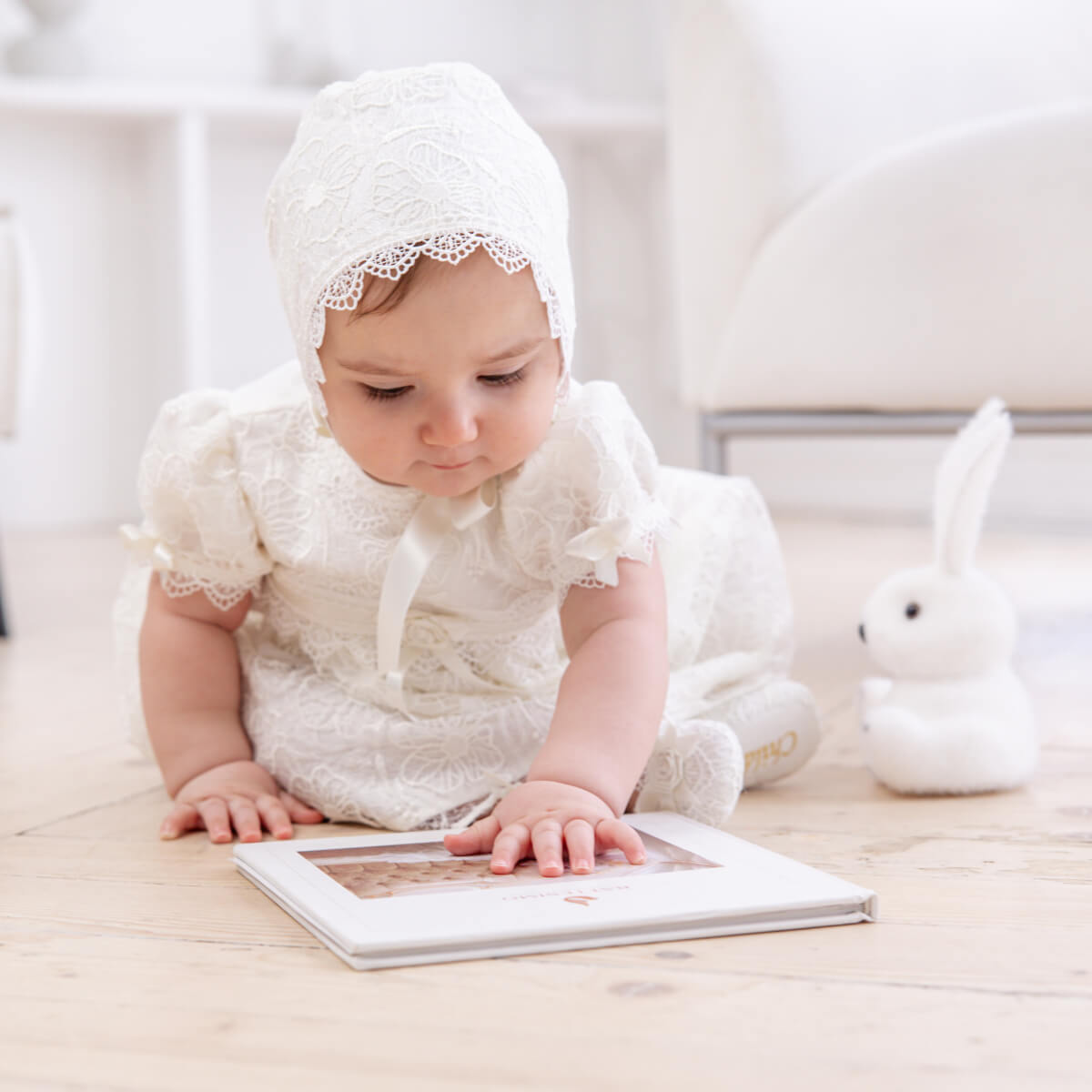 Baby in a Lace Baptism Dress and bonnet sitting on a wooden floor with a photo album.