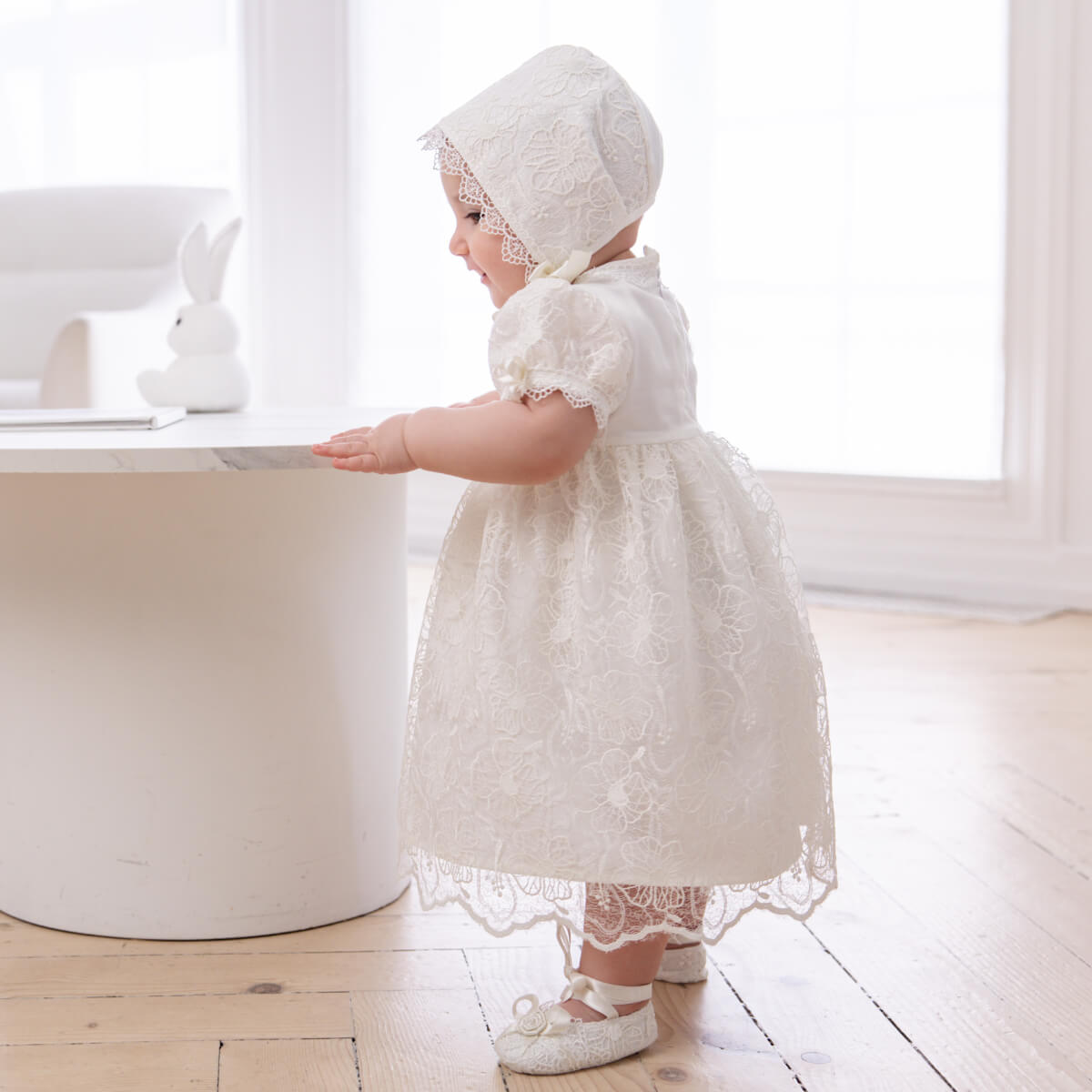 Baby in a Lace Baptism Dress and bonnet standing on a wooden floor.