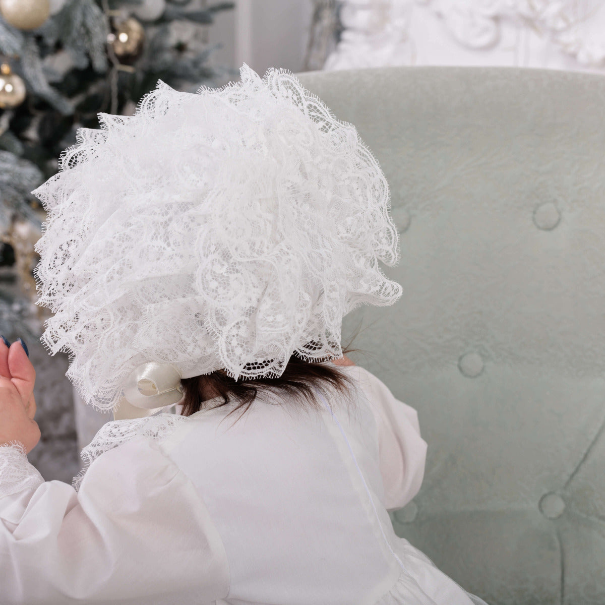 Person wearing a Lace Baptism Bonnet  sitting on a couch with a blurred decorative background