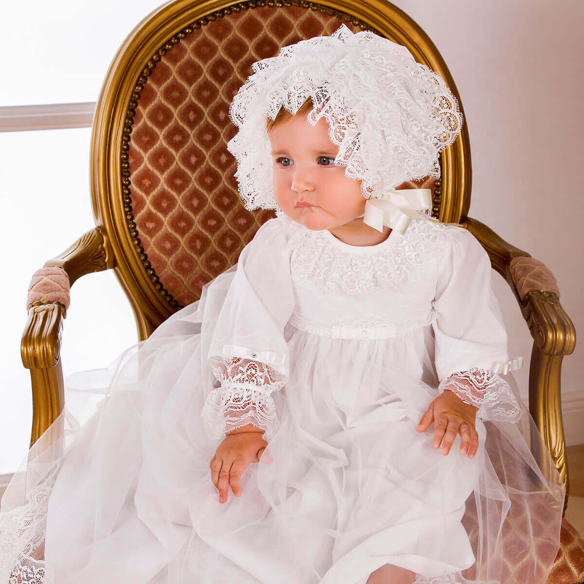 Baby in a Christening Gown for Girls sitting on a decorative chair.