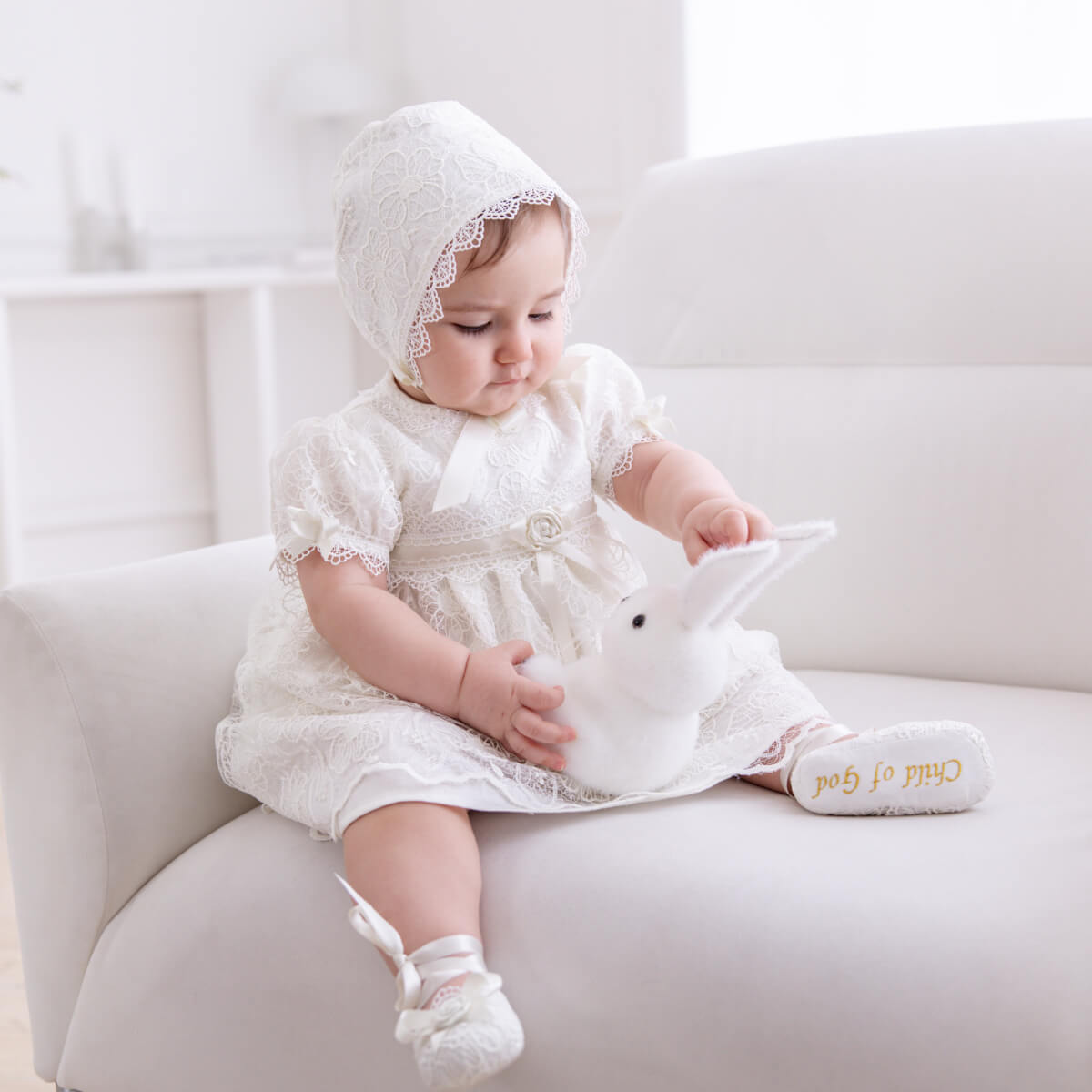 Baby in a white dress and Cotton Baptism Bonnet holding a white toy on a white couch.
