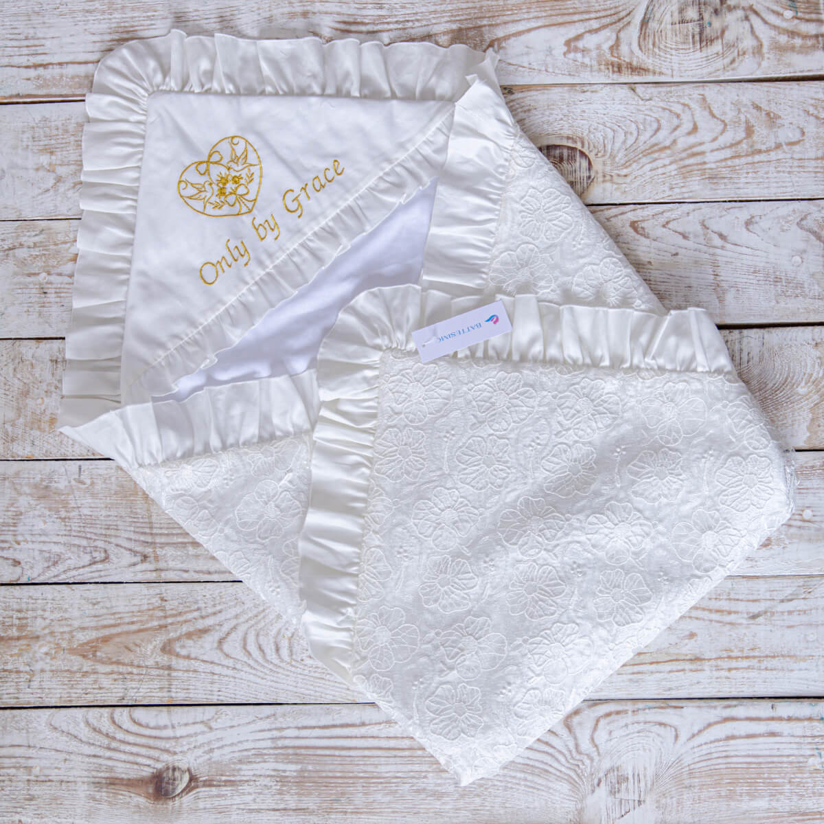 White baby blanket with ruffled edges and embroidered text on a wooden surface