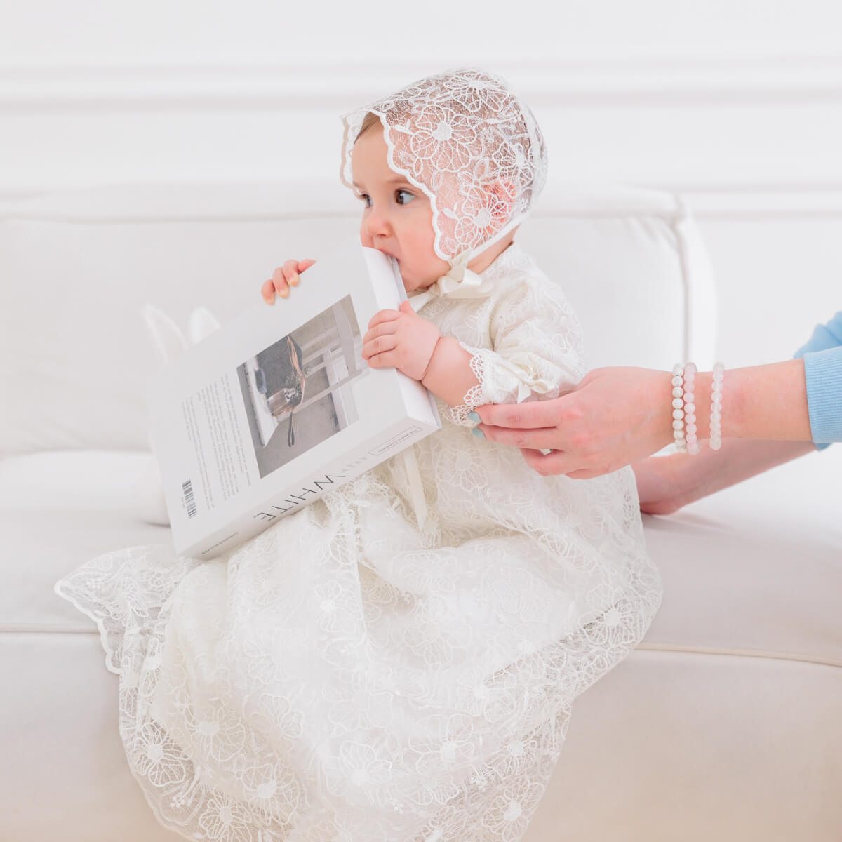 Baby in a Christening Gown for Girls holding a book, sitting on a white couch.