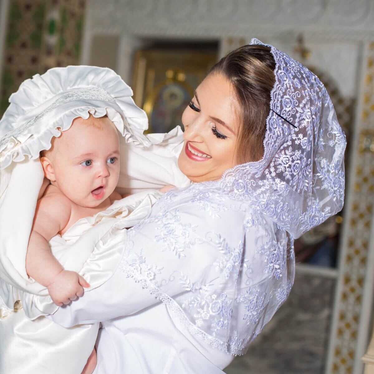 Woman in a white dress with a lace headscarf holding a baby also in a white dress with a Lace Caped Veil