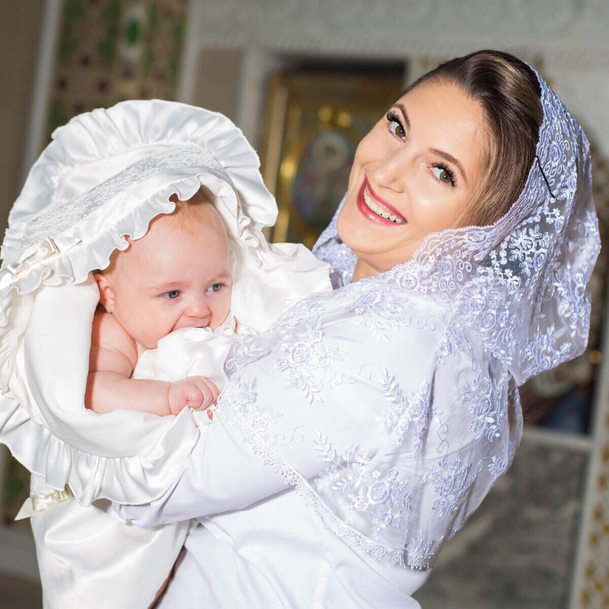 Woman in Lace Caped Veil holding a baby wrapped in white cloth with lace details, smiling.