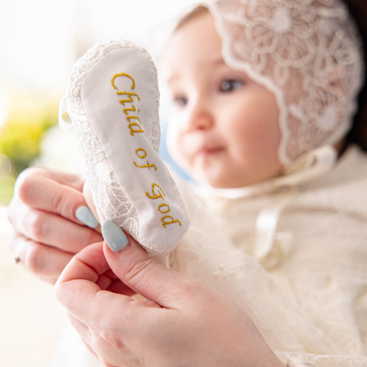 Baby's hand holding a white bootie with 'Child of God' text, wearing a lace bonnet.