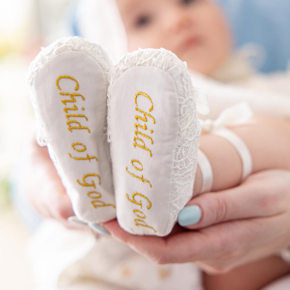 White booties with 'Child of God' embroidery held by a person, with a blurred background.