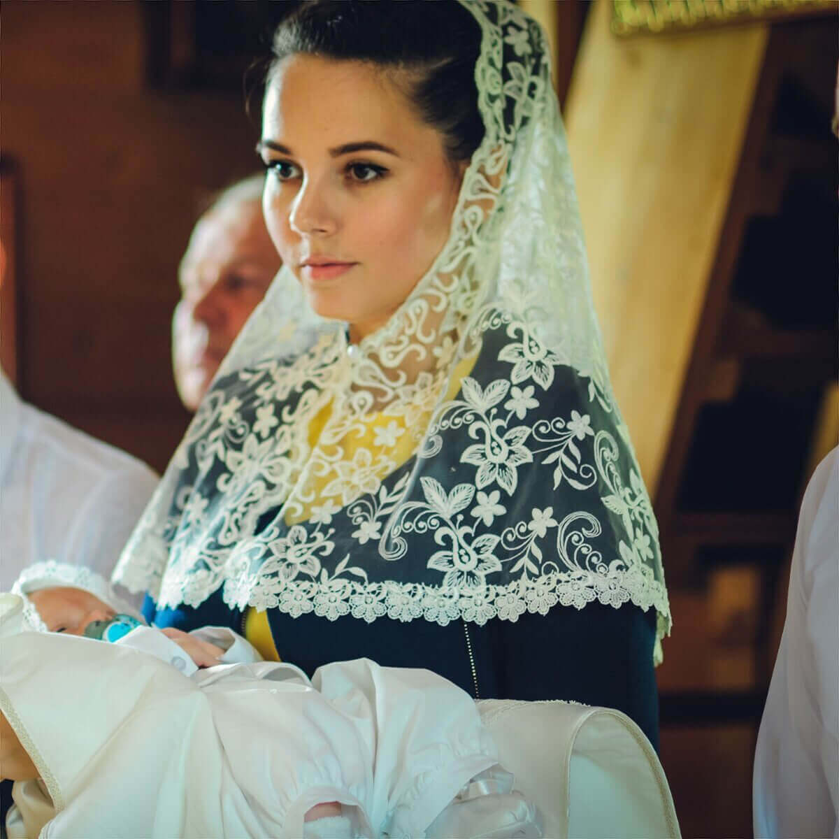 Woman wearing a Catholic Church Mantilla holding a baby in a warm indoor setting.
