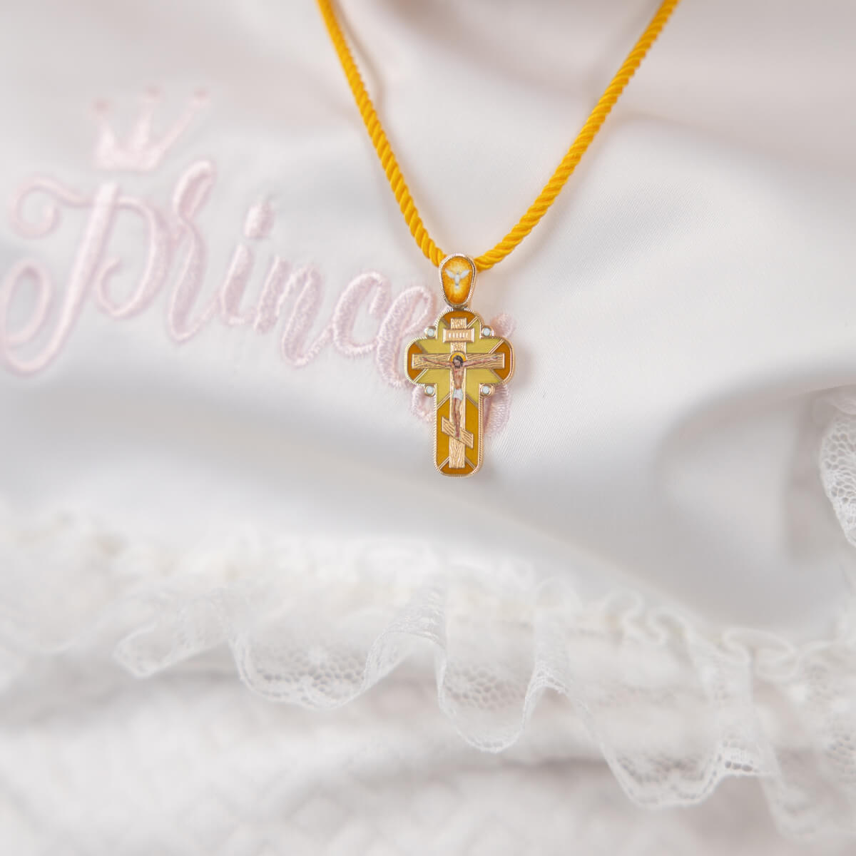 SAVIOR Baptism Cross – Handcrafted Gold Enamel Crucifix on a yellow string against a white lace background