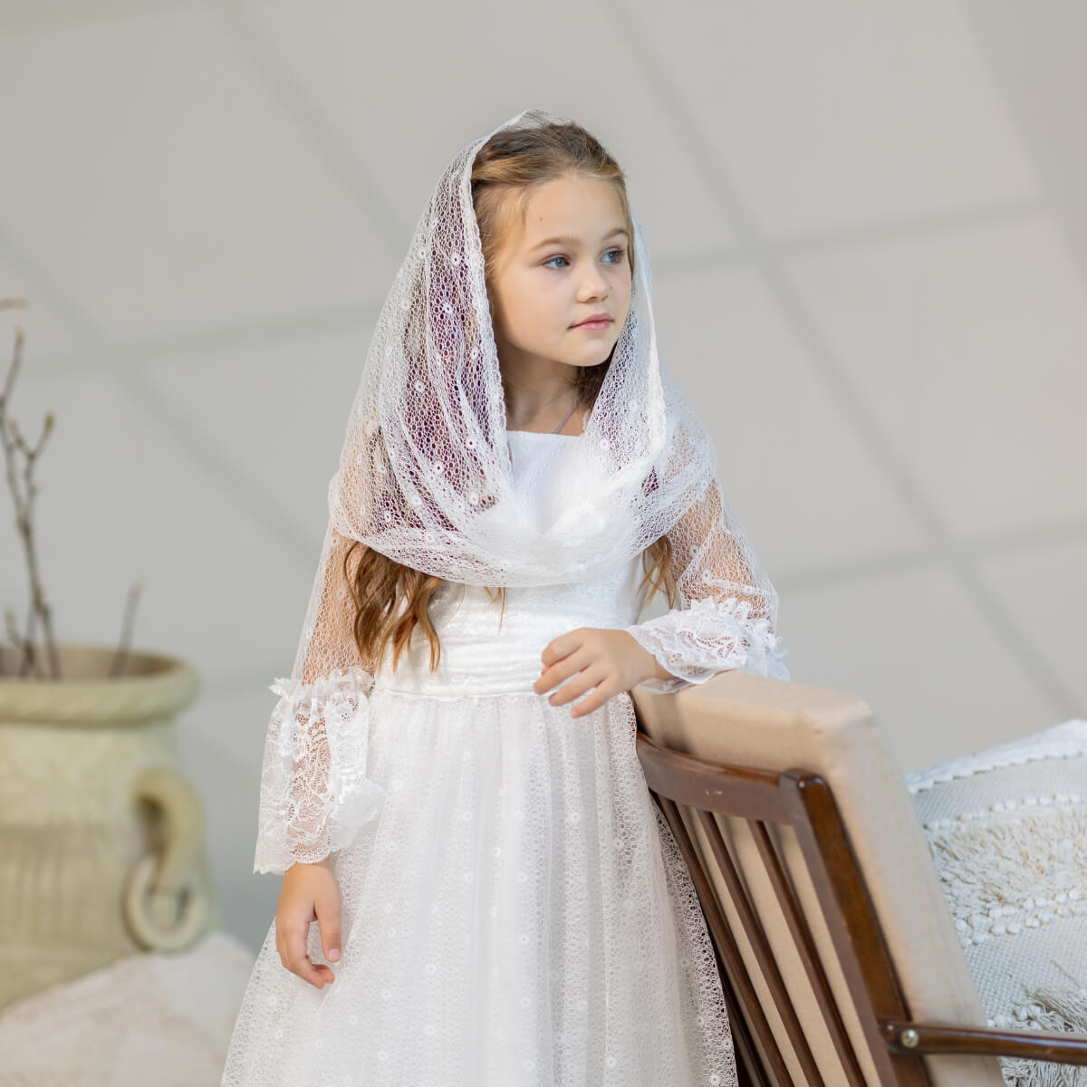 Young girl in a white lace dress with a First Communion Mantilla Veil against a neutral background