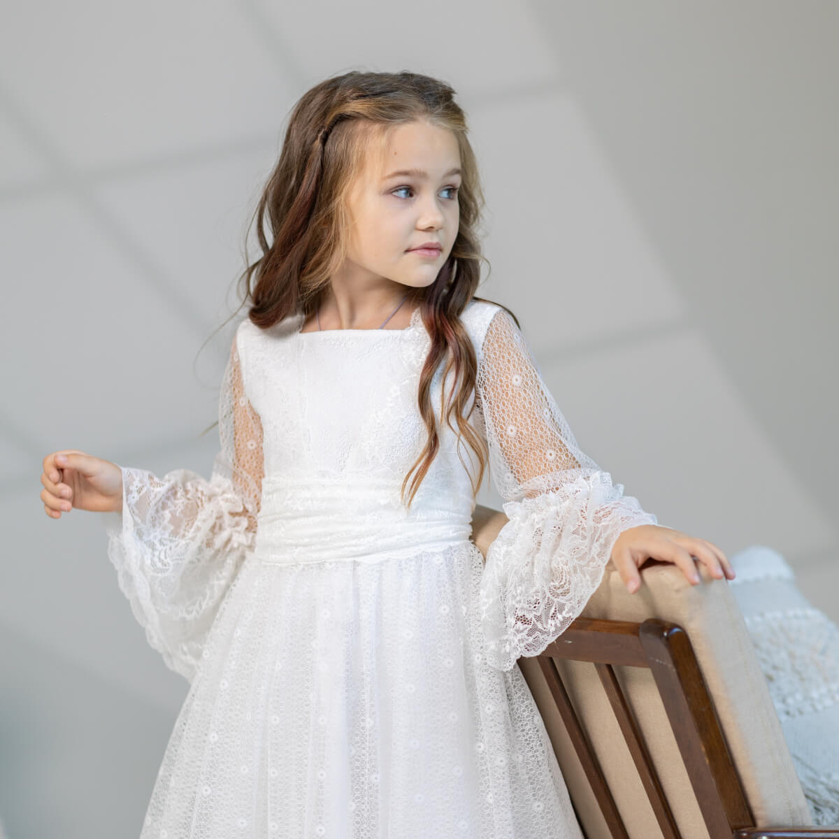 Young girl in a white lace dress sitting on a chair against a gray background