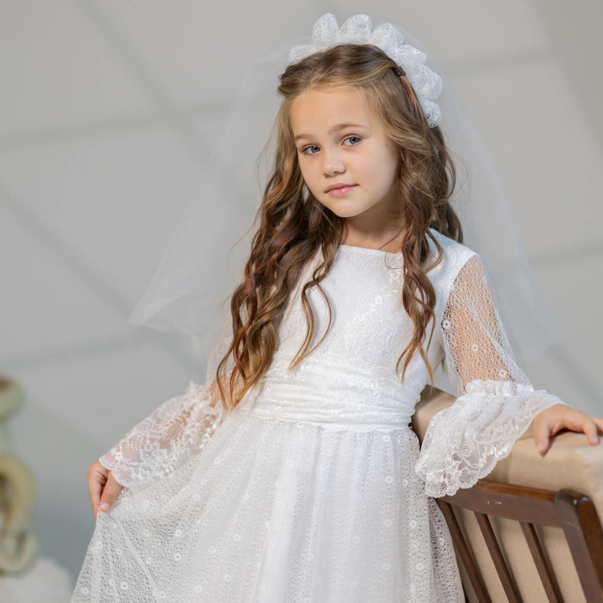 Young girl in a Elegant cotton communion dress with a tiara, sitting on a chair against a plain background