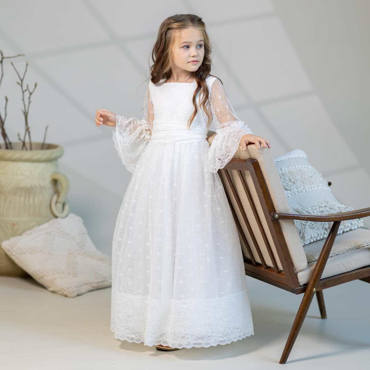 Young girl in a Elegant cotton communion dress standing in a room with a chair and decorative items.