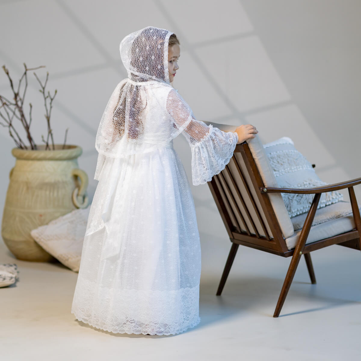 Person wearing a white lace dress and  First Communion Mantilla Veil standing next to a wooden chair with a textured wall background.