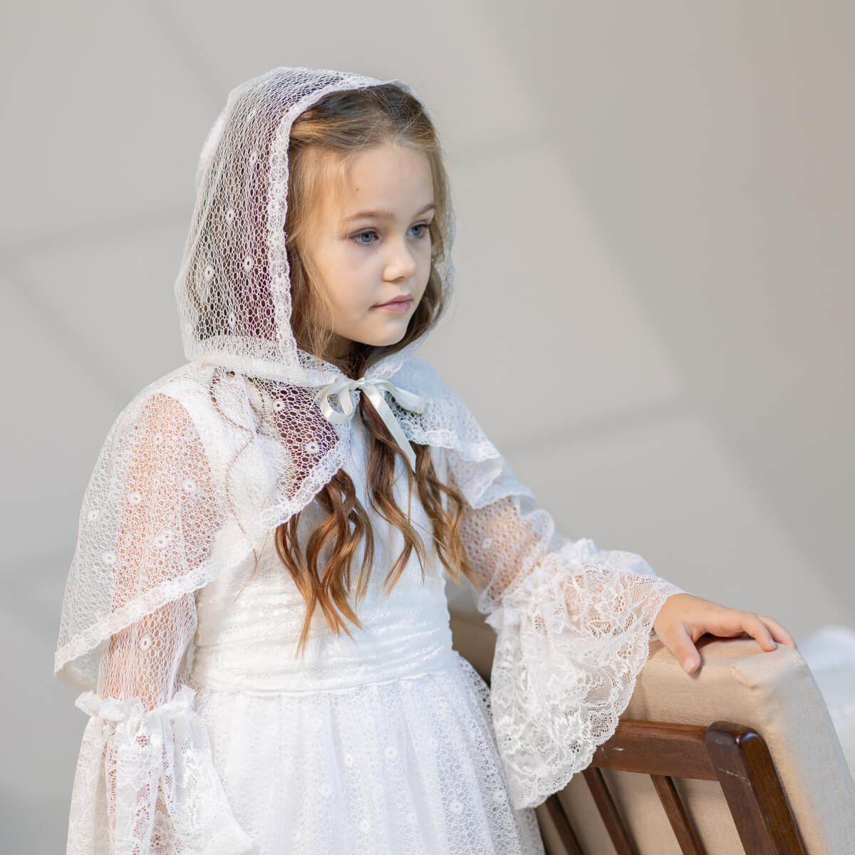 Young girl wearing a white lace dress and  First Communion Mantilla Veil against a plain background