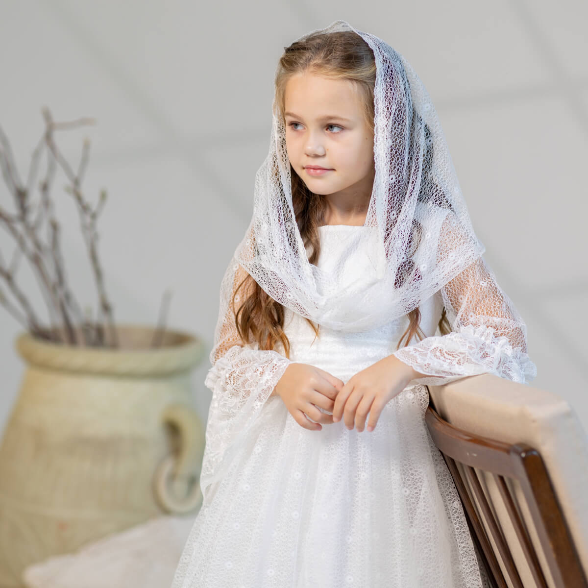 Young girl in a white lace dress and First Communion Mantilla Veil sitting on a wooden stool against a neutral background.