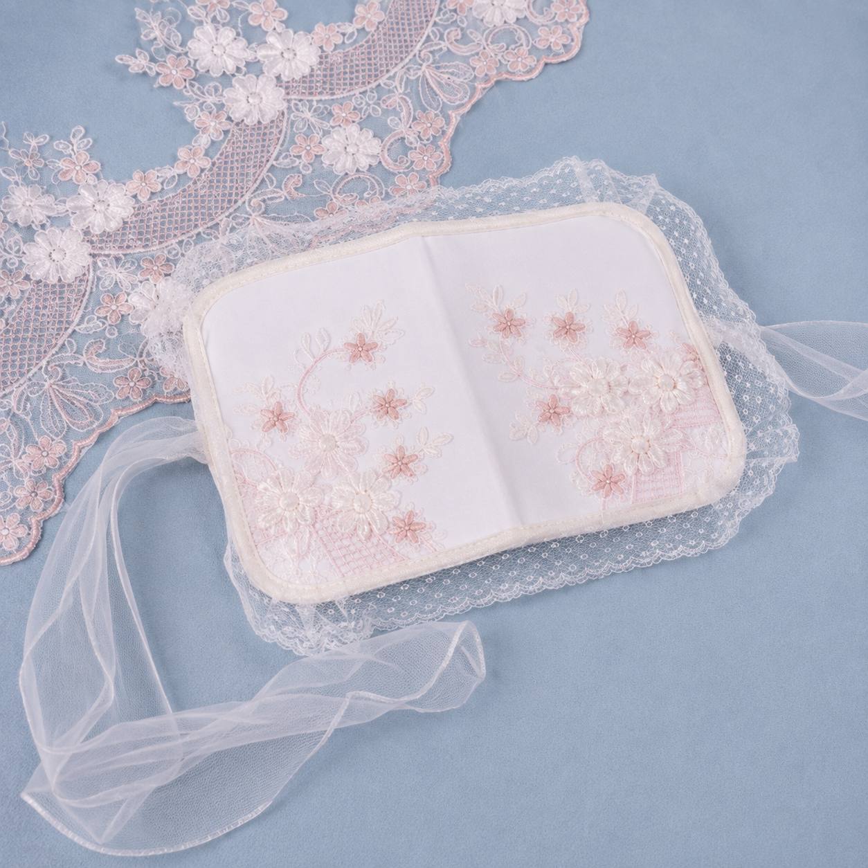 A small white embroidered pouch or book-shaped accessory with floral stitching, placed on a light blue background with lace and sheer tulle fabric props.