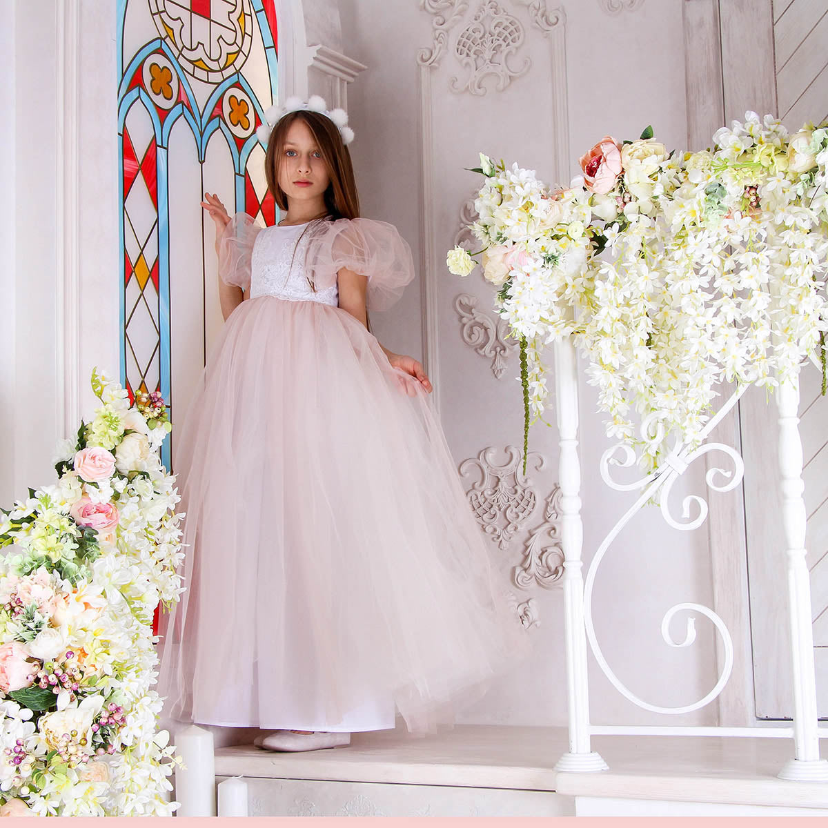 Woman in a white dress standing in a decorated room with flowers and a stained glass window.