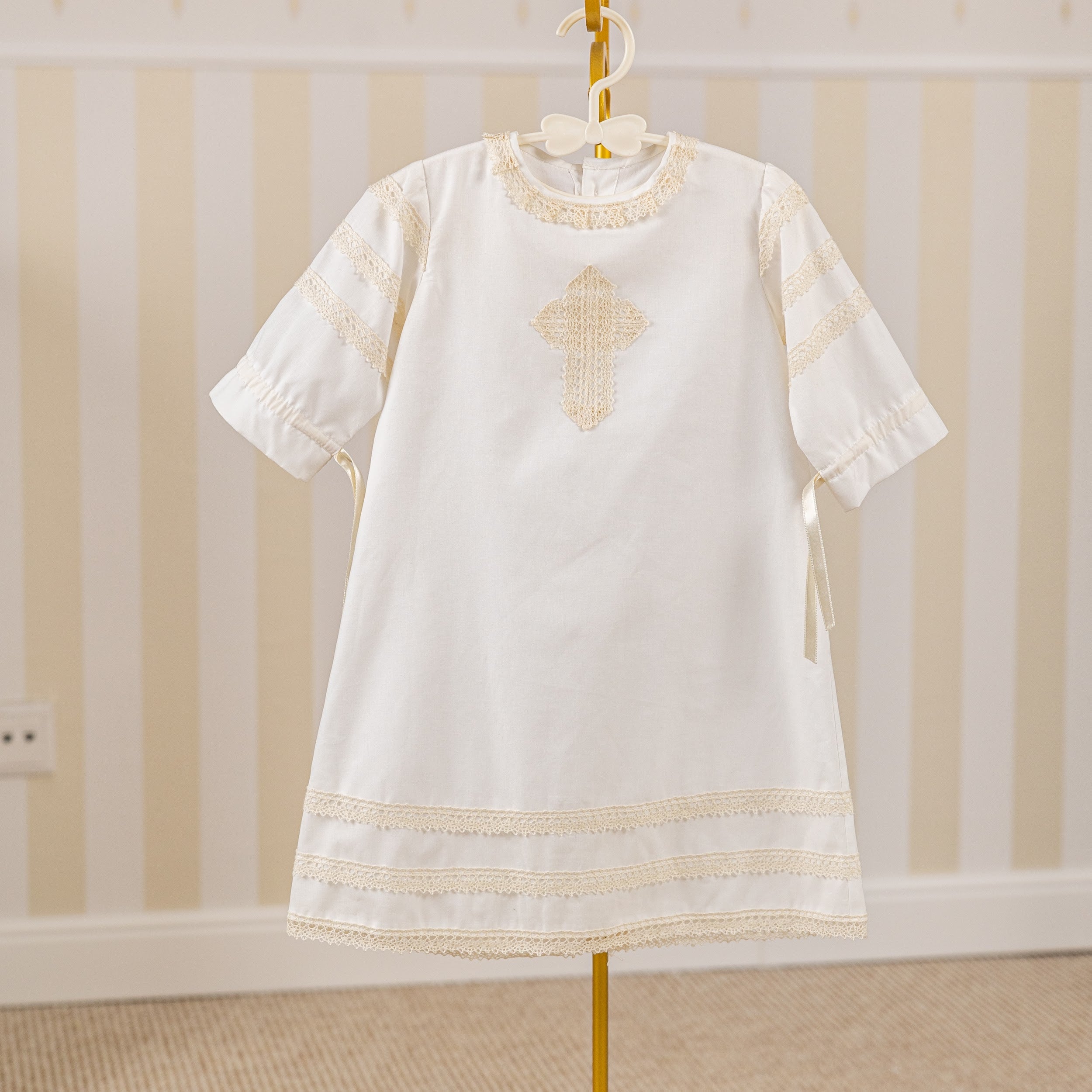  Baptism Gown with gold embroidery on a hanger against a striped wall.