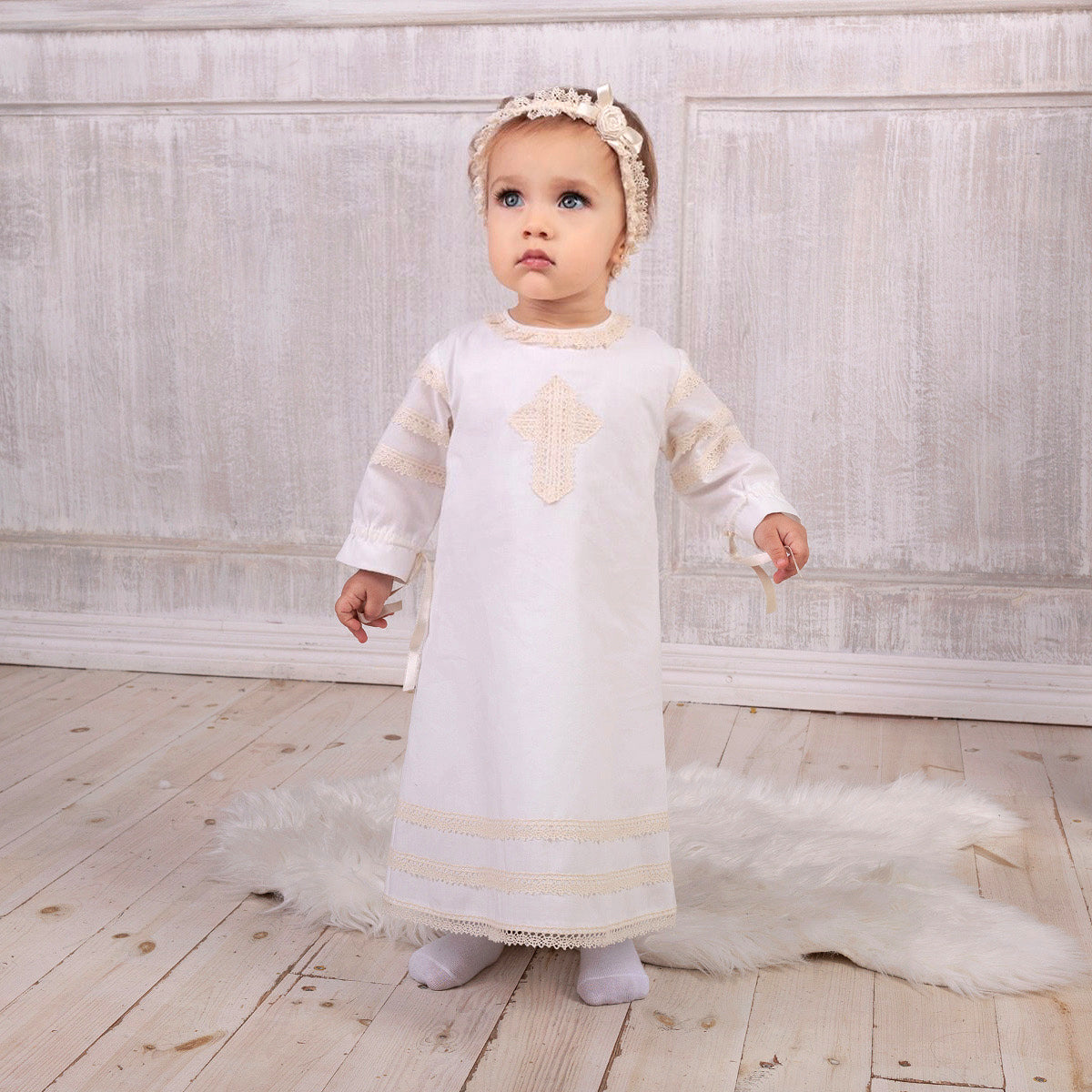 Baby in a  Baptism Gown with a cross design, standing on a wooden floor.