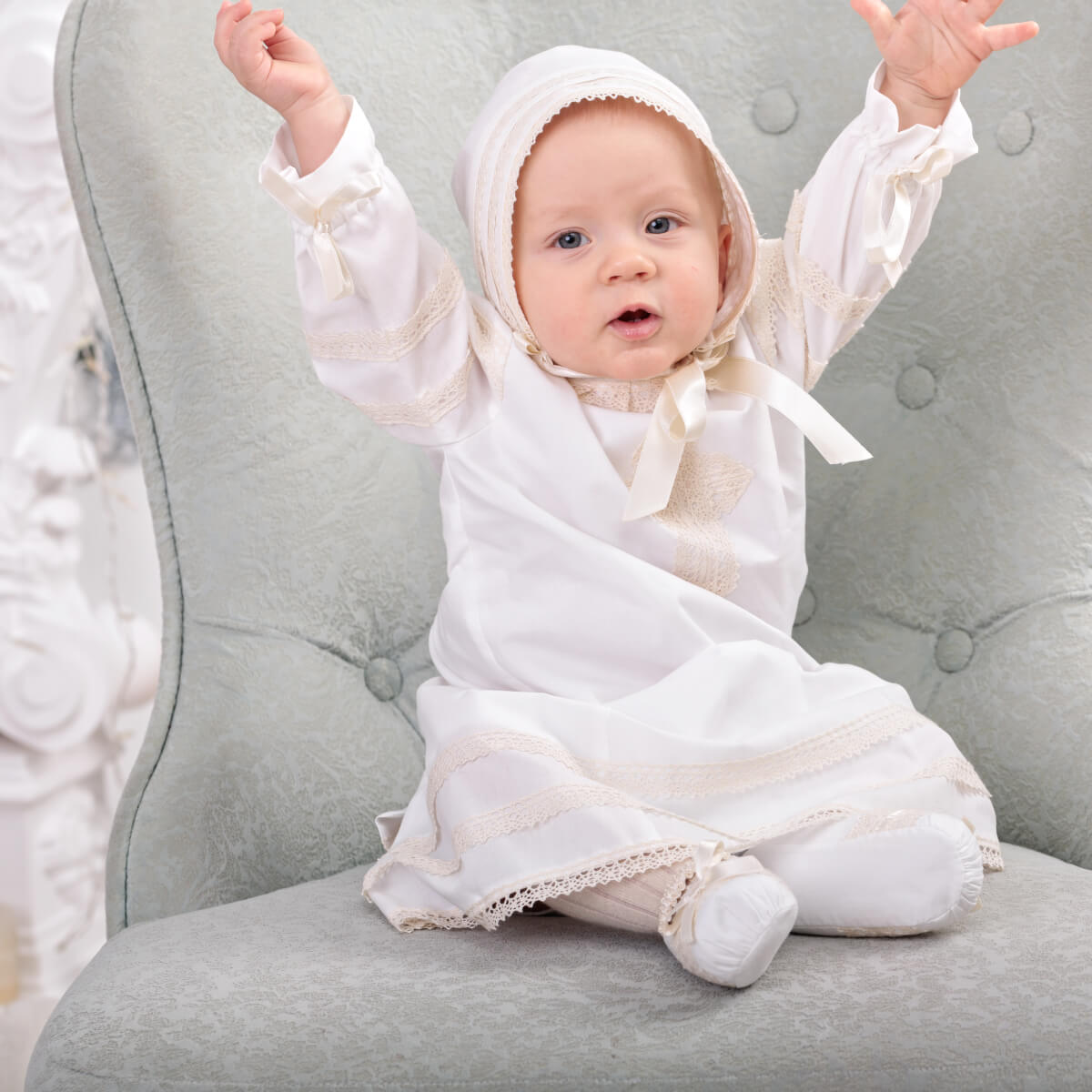 Baby in a  Baptism Gown with a hood sitting on a gray chair.