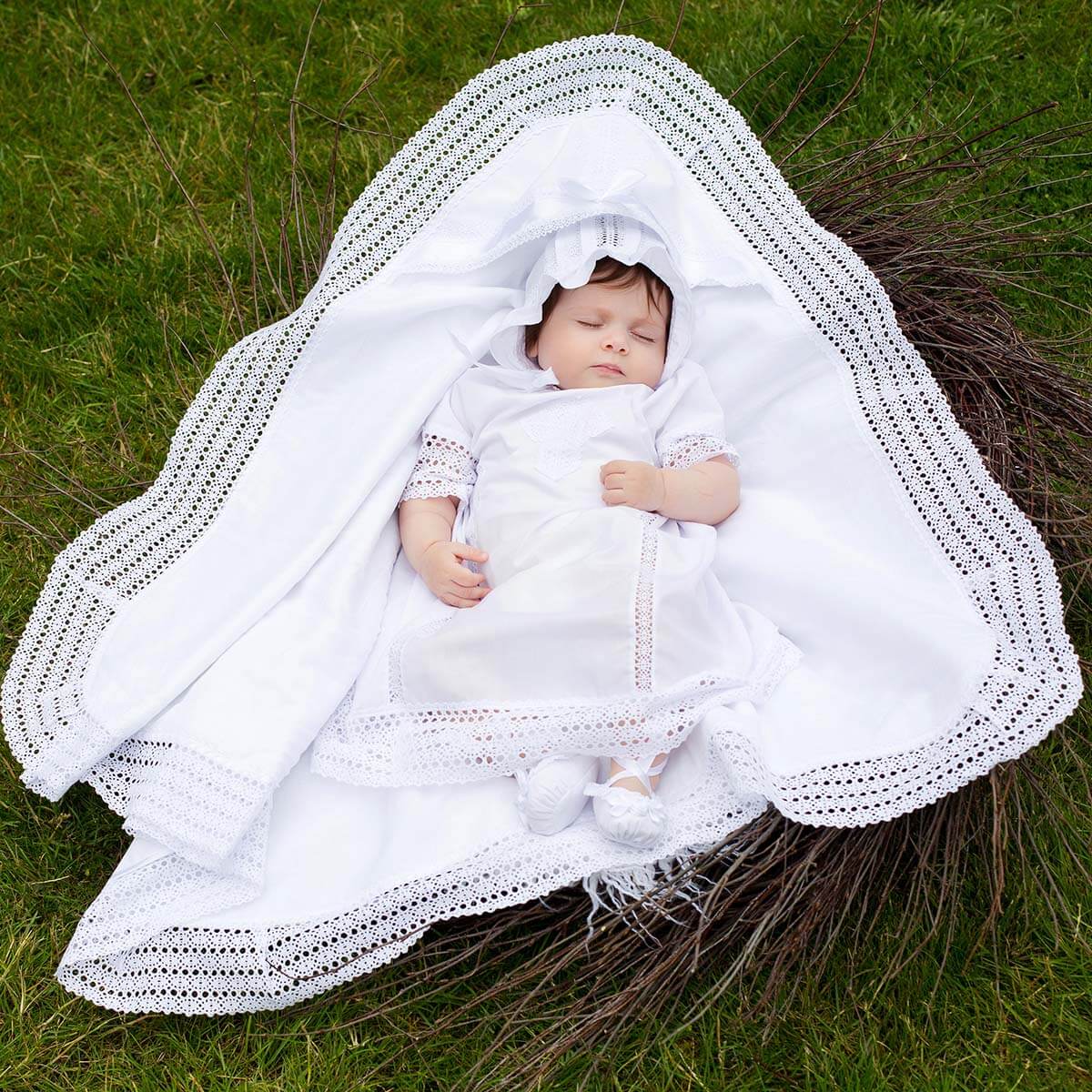 Baby wrapped in a white blanket with lace details, lying on grass.