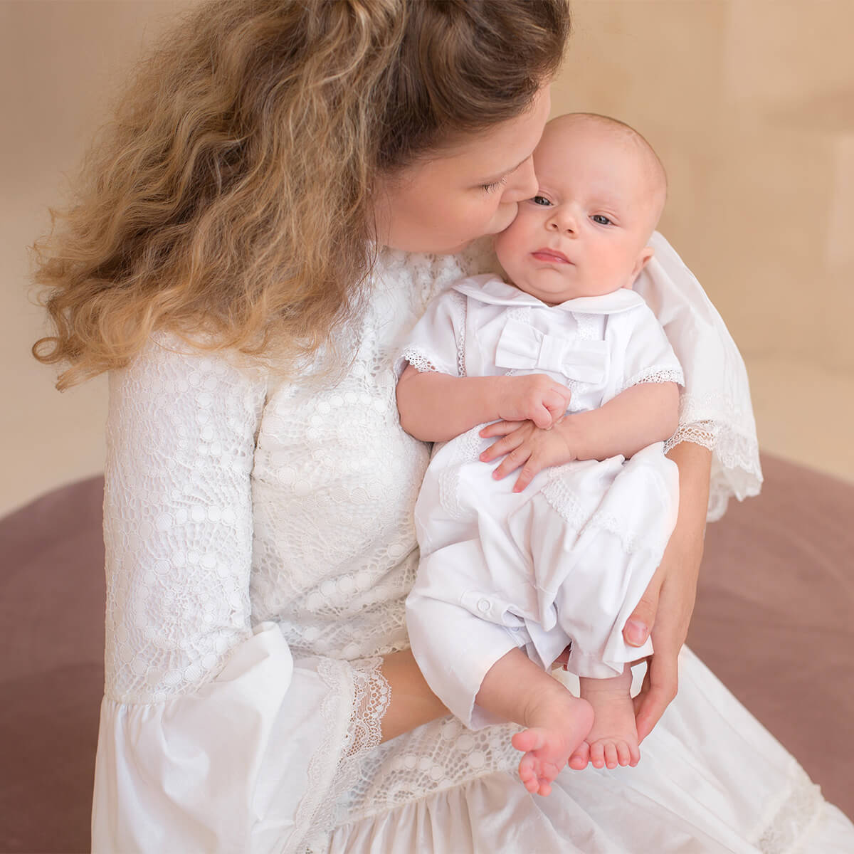 Woman in a white dress holding a baby in Baby Boy Christening Romper a similar outfit against a beige background