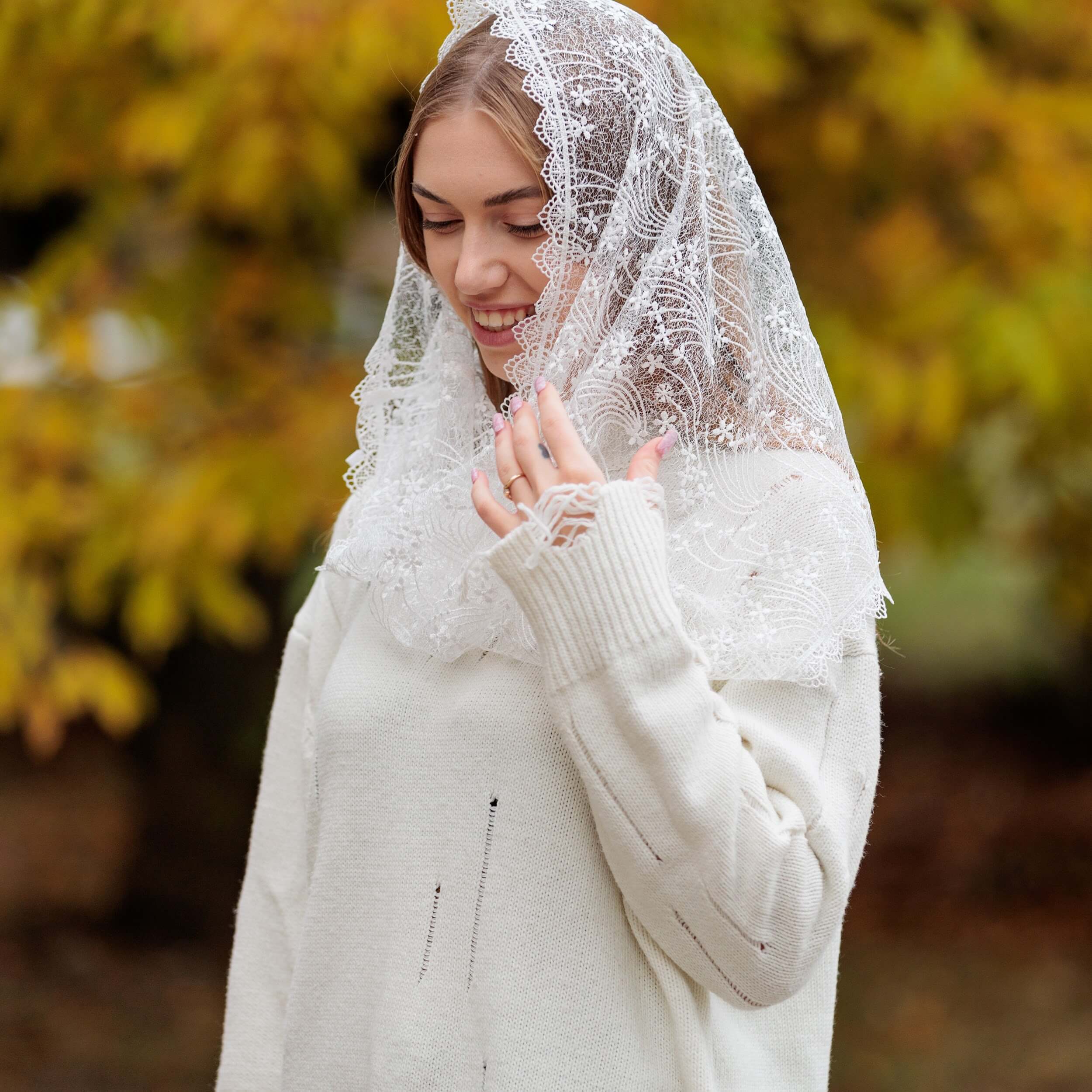 Woman wearing a white Catholic Infinity Veil outdoors with blurred autumn trees in the background