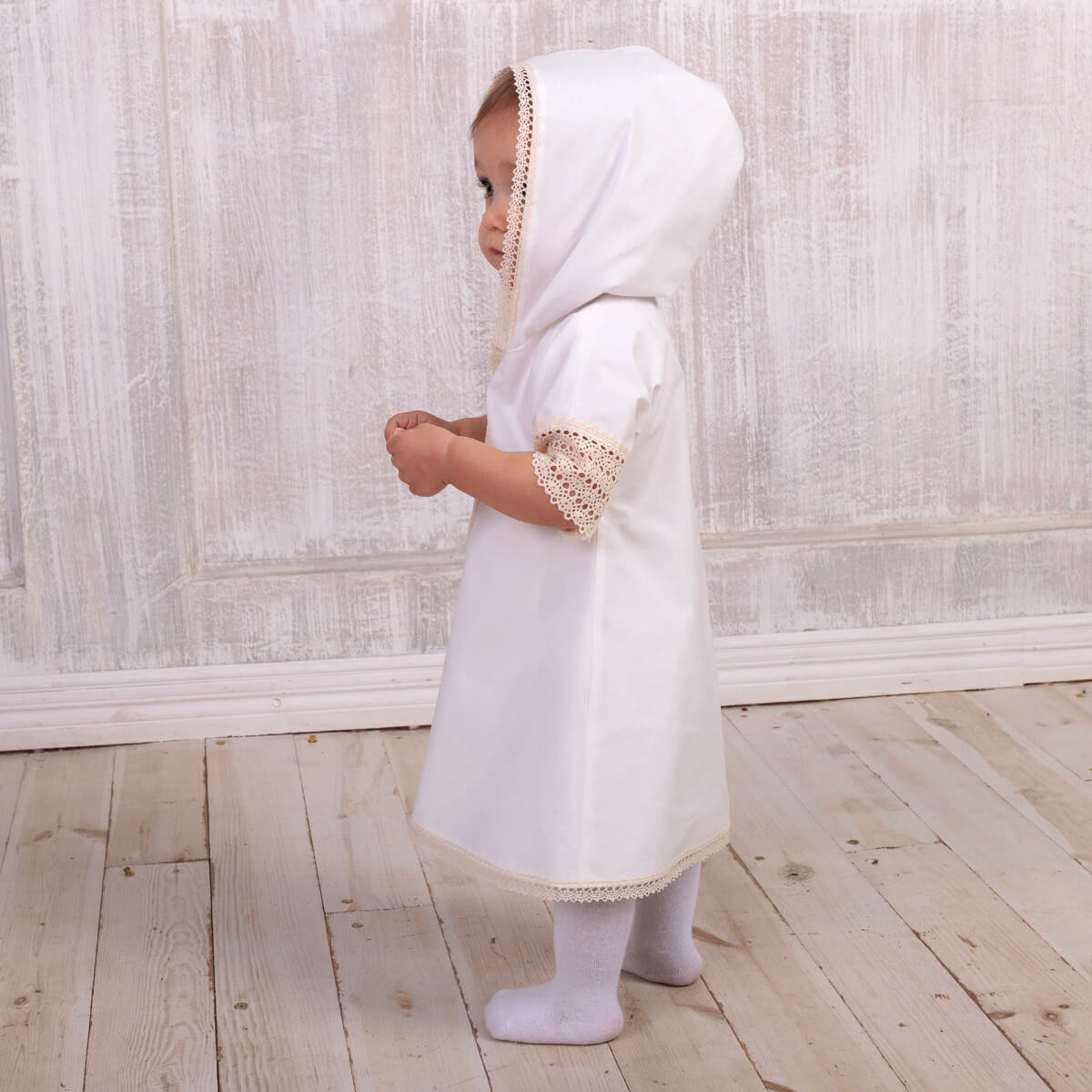 Child wearing a Baptism Gown for Boy with a hood in a room with wooden flooring and a light-colored wall.