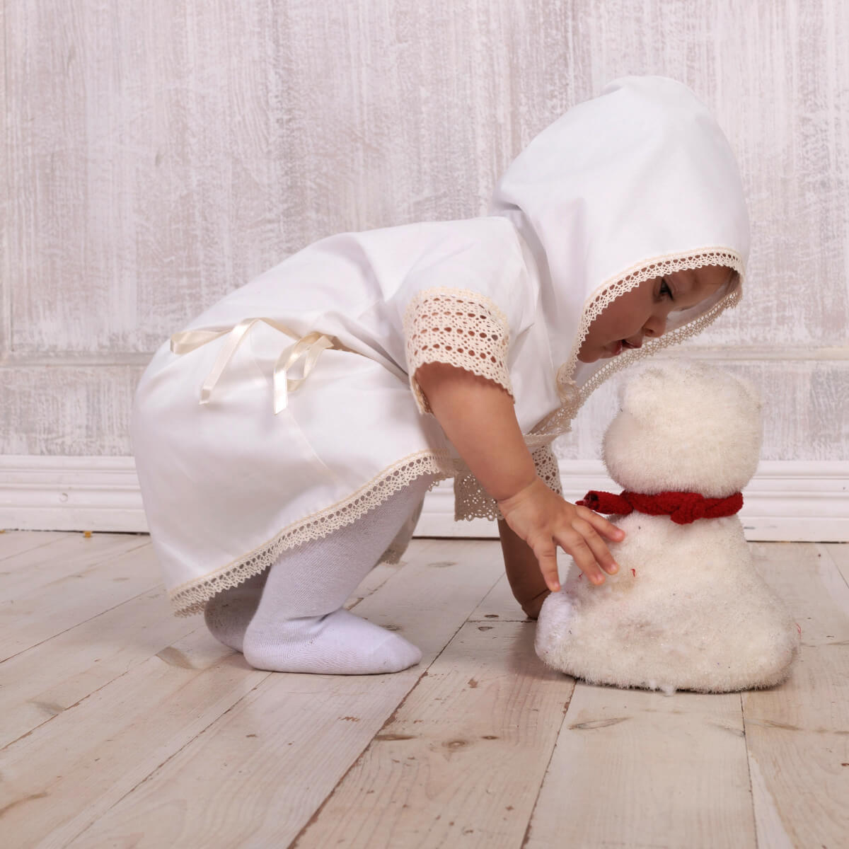 Child in a white Baptism Gown for Boy playing with a small snowman on a wooden floor.