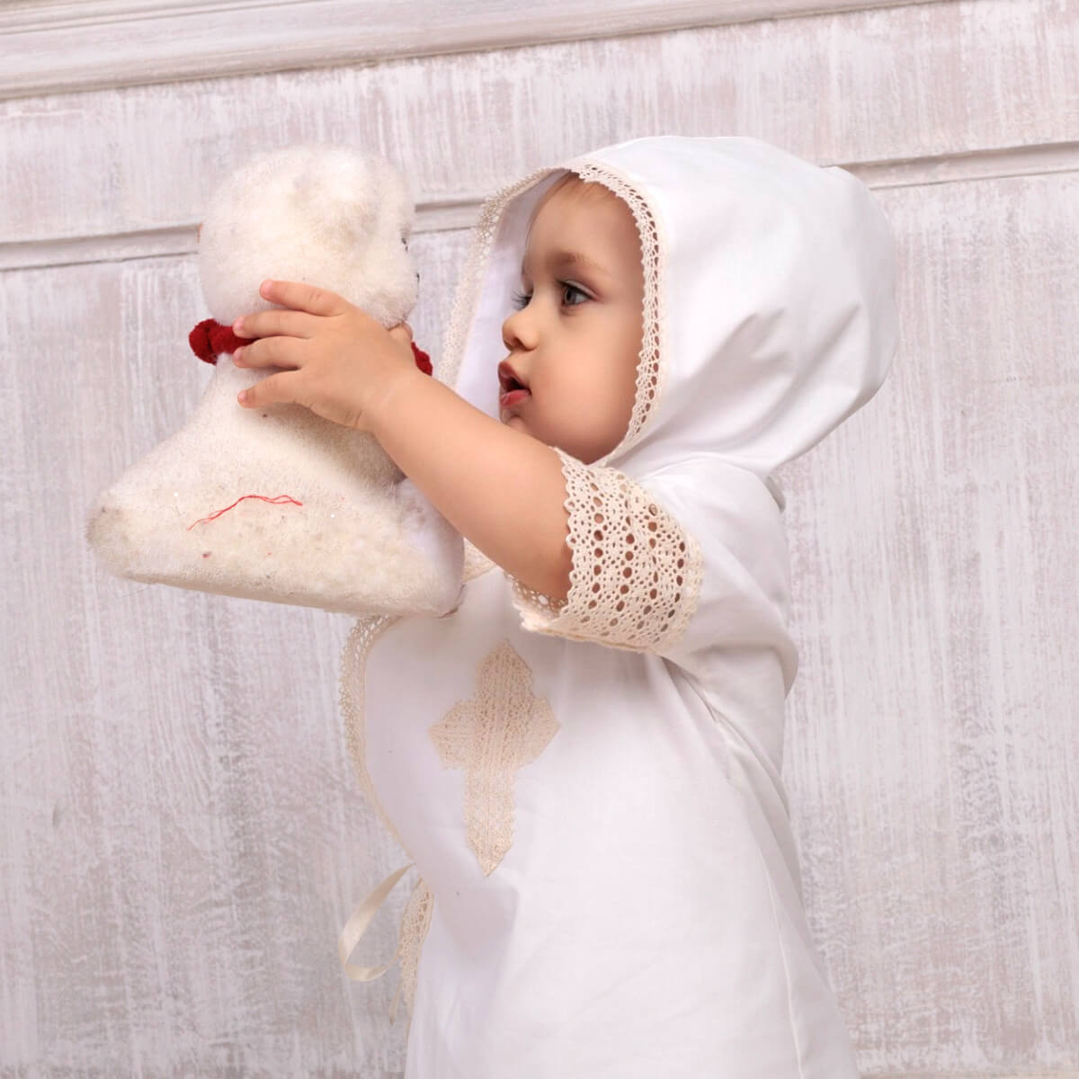 Baby in a Baptism Gown for Boy with a hood holding a white boot with red details against a light wooden background